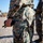 Close-up photograph of a military personnel wearing digital camouflage uniform, viewed from behind at torso level. The individual is holding what appears to be a tactical pen or small tool while wearing tactical gloves and a wristwatch. A second uniformed individual is visible in the blurred background, along with a military vehicle.