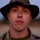 A close-up portrait of a young male service member wearing MARPAT (Marine pattern) camouflage utility uniform and a matching camouflage boonie hat. The subject is looking directly at the camera with a neutral expression. The background is blurred, suggesting an outdoor military training or field environment.