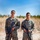 Two U.S. Marines in MARPAT woodland digital camouflage uniforms stand at ease in an outdoor field environment. Both are holding rifles and wearing name tapes, with the right Marine's tape reading 'U.S. MARINES' clearly visible. The setting appears to be a training range or field exercise area with dry grass and a tree line in the background.