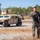 A military service member in digital camouflage uniform stands in the foreground of an arid training or operational area, holding a rifle at a relaxed carry position. Behind him, a tan-colored HMMWV (Humvee) military vehicle is parked on a dirt road, with sparse vegetation and trees visible in the background. The image appears to be a professional or documentary-style military photograph taken in a dry, open terrain environment.