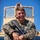 A U.S. Marine in MARPAT woodland digital camouflage uniform is seated on top of a military vehicle, laughing broadly. He is holding an M16-style rifle across his body in a relaxed, informal pose. The background features a tan military vehicle and a clear blue sky.