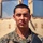 A young male service member in U.S. Marine Corps MARPAT woodland/desert digital camouflage uniform is photographed in close-up portrait style. He is holding what appears to be a rifle or weapon system and is positioned in front of a tan-colored military vehicle, likely a HMMWV (Humvee). The image is a sharp, well-lit outdoor photograph taken in bright daylight.