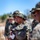 A group of military personnel in digital camouflage uniforms and boonie hats are gathered outdoors, likely during a field training exercise. The central figure is holding a small notepad and a rifle, with a helmet secured to their pack. The setting appears to be a dry, open training area under clear blue skies.