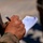 A person wearing military camouflage (MARPAT pattern, consistent with U.S. Marines) is writing notes on a small lined notepad while holding a pen. A rifle barrel is visible in the upper left, and what appears to be an American flag is partially visible in the lower right. The image is taken in an outdoor setting with a blurred background.
