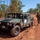 A military HMMWV (Humvee) is parked on a red dirt road in what appears to be a dry tropical or savanna environment. A soldier in camouflage uniform carrying a weapon is walking alongside the vehicle. The setting and vegetation are consistent with northern Australia, likely during a military exercise.