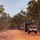 A convoy of military vehicles, appearing to be US-style HMMWVs (Humvees), travels along a red dirt road through sparse eucalyptus woodland. The terrain and vegetation are consistent with Australian outback or tropical savanna environment. The vehicles are traveling in a single-file formation with headlights on.