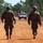 Two soldiers in camouflage uniforms walk along a red dirt road toward a tan/desert-colored Humvee (HMMWV) with a gunner positioned in the turret. The scene appears to be a military patrol or training exercise in an arid, eucalyptus-tree-lined environment consistent with northern Australia. The image is taken from behind the soldiers, creating a perspective shot down the road toward the vehicle.