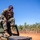 A military service member in MARPAT (Marine pattern) camouflage uniform and tactical vest sits atop a military vehicle or equipment case, speaking into a corded radio handset. The setting appears to be a dry, arid training area with red dirt terrain and sparse eucalyptus-type vegetation, consistent with northern Australia. The image is well-lit under clear blue skies, suggesting a field exercise or operational deployment.