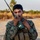 A young male service member in U.S. Marine Corps MARPAT digital camouflage uniform is holding a military tactical radio (appears to be a PRC-type handheld radio) up to his mouth, likely transmitting or receiving a communication. He is wearing a tactical vest/plate carrier with pouches and is standing on an open dirt road or airstrip. The background shows dry grassland and sparse trees under a hazy sky, suggesting a warm, arid environment possibly during golden hour.