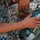 A person wearing US Marine Corps MARPAT digital camouflage uniform is handling a military tactical radio, appearing to operate or configure it. The device is an olive-drab green military-grade radio with a keypad, small display screen, and multiple connectors. The setting appears to be an outdoor gravel or rocky surface.
