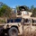 A tan/desert-camouflage HMMWV (Humvee) military vehicle is positioned on a dirt track in a dry, scrubby woodland environment. A soldier wearing a camouflage hat is visible manning the turret position atop the vehicle, which is equipped with a weapon system and camouflage netting. The setting and vegetation suggest a hot, arid training or operational environment, possibly in northern Australia.