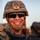 A smiling military service member is photographed in a close-up portrait shot. The individual is wearing digital camouflage (MARPAT pattern consistent with U.S. Marine Corps), a combat helmet with a mounted device, ballistic sunglasses, and body armor. The warm golden lighting suggests the photo was taken during sunrise or sunset in an arid environment.