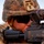 A close-up photograph of a military service member in full combat gear aiming down the sights of a rifle. The individual is wearing MARPAT (Marine pattern) digital camouflage, a combat helmet with a tan accessory mount, and dark tactical sunglasses. The image is tightly framed, emphasizing the weapon optic and the soldier's focused posture.