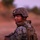 A U.S. Marine in full combat gear is photographed at dusk or dawn in an arid, desert-like environment. The service member is looking back over their shoulder toward the camera while holding what appears to be a small device. The warm reddish-orange ambient light suggests either sunrise or sunset conditions.