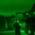 A night vision (NVG) captured image showing military personnel operating in a vehicle staging area during nighttime hours. The green monochromatic imagery is characteristic of Generation 2 or 3 image intensification night vision equipment. At least two personnel are visible among what appear to be military tactical wheeled vehicles.