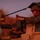 A U.S. Marine in MARPAT camouflage uniform and combat helmet is positioned in the turret of a light-colored military vehicle, appearing to load or operate a crew-served weapon system. The image is taken during low-light conditions at dusk or dawn, casting a strong reddish-orange ambient light across the scene. Camouflage netting is visible draped over the vehicle's turret area.