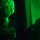 A low-light image illuminated entirely by green light, showing at least one person wearing what appears to be a helmet or head covering. The scene is very dark with the green light source casting strong shadows on a wall to the right. The image has a tactical or night-operation aesthetic.
