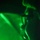 A close-up image captured under intense green lighting, showing what appears to be a metallic chain or hook mechanism against a dark background. The green illumination creates strong contrast between lit and shadowed areas, giving the image a dramatic, industrial or tactical appearance. The shallow depth of field and low-light conditions make precise identification of all elements difficult.