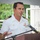 A naval officer in white dress uniform is speaking at a podium with a microphone. He is gesturing with his left hand while addressing an audience at what appears to be an outdoor ceremony. The setting includes a covered area with trees visible in the background.