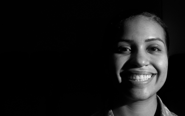 A black and white portrait photograph of a female U.S. Air Force service member wearing Airman Battle Uniform (ABU) digital camouflage. She is smiling broadly and is lit dramatically against a dark background. Partial text on her uniform reads 'U.S. AIRFORCE' and a partial name tape is visible.