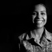 A black and white portrait photograph of a female U.S. Air Force service member wearing Airman Battle Uniform (ABU) digital camouflage. She is smiling broadly and is lit dramatically against a dark background. Partial text on her uniform reads 'U.S. AIRFORCE' and a partial name tape is visible.