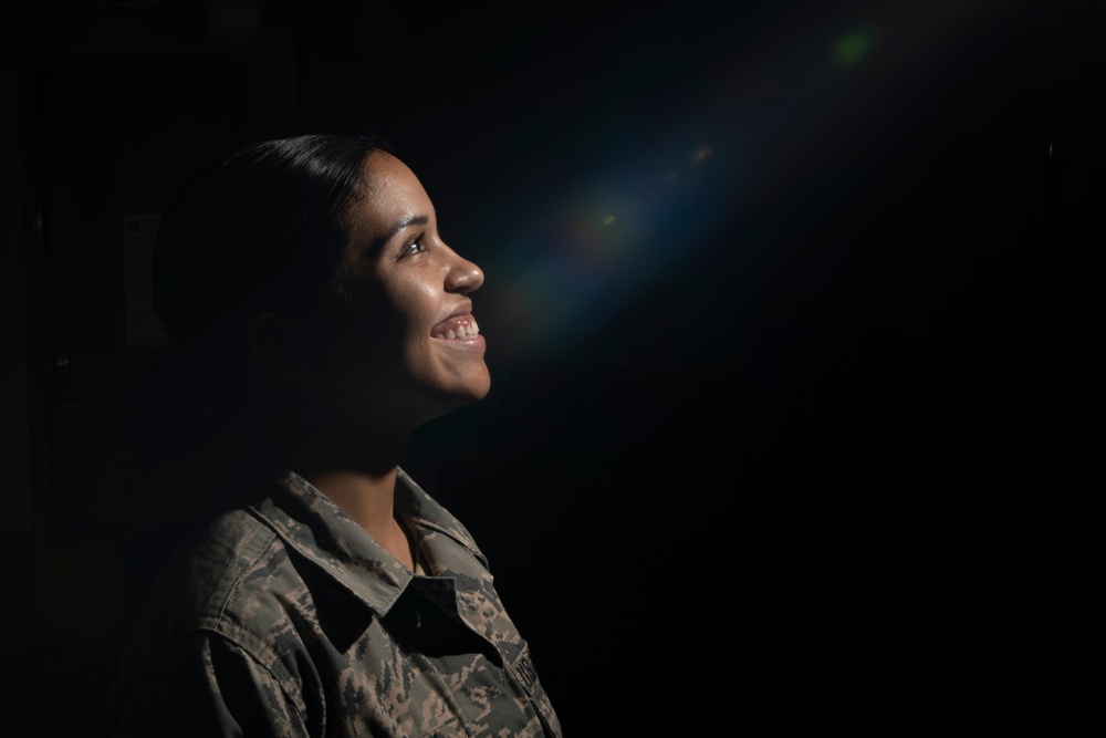 A woman in military camouflage uniform is photographed in a dramatic low-key portrait style against a dark background. She is smiling and looking upward, illuminated by a focused light source that creates strong contrast. A lens flare is visible in the upper right portion of the frame, adding an artistic element to the composition.