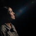A woman in military camouflage uniform is photographed in a dramatic low-key portrait style against a dark background. She is smiling and looking upward, illuminated by a focused light source that creates strong contrast. A lens flare is visible in the upper right portion of the frame, adding an artistic element to the composition.