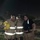 Three women in firefighter turnout gear pose together outdoors at night, smiling for the camera. Two are wearing full tan/gold bunker gear with reflective strips, while the center person also wears a firefighter helmet with a name tag reading 'K. SOTO' and 'PHARR'. The photo appears to be taken at a training exercise or active scene, with a chain-link fence visible in the background.