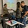 A military service member in camouflage uniform is conducting an anti-bullying and safety presentation in a classroom setting. Students are actively engaged, with some signing a black poster board while others interact with the presenter. A projected slideshow displays a message about safety, respect, and bullying prevention.