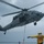 A U.S. Navy helicopter, numbered 616, hovers low over a ship's deck during what appears to be a hoist or vertical replenishment operation. A deck crew member in a yellow jersey and cranial helmet directs the aircraft with outstretched arms. The overcast sky and dynamic rotor blur suggest active flight operations at sea.