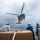A military helicopter is conducting a vertical replenishment (VERTREP) operation above the deck of a naval vessel, lifting cargo via a sling load. Three crew members in protective gear observe and manage the operation from the ship's deck. The vest of one crew member is visibly marked 'USS STERETT DDG 104', identifying the vessel.