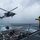 A military helicopter, appearing to be an MH-60 Seahawk, is conducting a vertical replenishment (VERTREP) operation over the flight deck of a naval warship in rough, overcast sea conditions. A deck crew member in a yellow jersey and helmet is directing the helicopter from the ship's railing while a cargo net with supplies is suspended beneath the aircraft. The operation is taking place in challenging weather with choppy seas and heavy cloud cover.