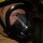 A close-up photograph of a firefighter wearing full protective gear, including a tan/gold turnout coat, a firefighting helmet with yellow trim, and a black full-face SCBA (Self-Contained Breathing Apparatus) mask. The individual's eyes are visible through the clear visor, appearing alert and looking upward. The image is taken in a dark environment, suggesting an active or training firefighting scenario.