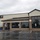 The image shows a military commissary building, identifiable by the 'COMMISSARY' sign on the facade, likely located on a U.S. military installation. The building features a modern commercial design with brick accents, large dark-tinted windows, and a metal standing-seam roof. The parking lot is wet from recent rain, and the area appears largely empty.
