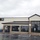 The image shows the exterior of a military commissary (grocery store) on a rainy, overcast day. The building features brick and stucco construction with a dark metal roof, large tinted windows, and a prominent 'COMMISSARY' sign with a logo. The wet parking lot in the foreground includes multiple ADA-accessible handicapped parking spaces.