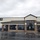 The image shows the exterior of a military commissary (grocery store) on a rainy, overcast day. The building features brick and stucco construction with a dark metal standing-seam roof and large tinted windows. The parking lot is wet and includes clearly marked handicapped accessible parking spaces near the entrance.