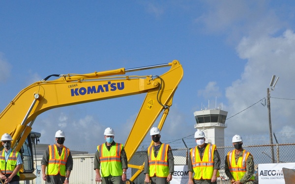 A groundbreaking ceremony is taking place at what appears to be a military airfield, with six individuals wearing hard hats, high-visibility vests, and face masks holding ceremonial gold shovels. A large yellow Komatsu excavator is prominently positioned behind the group, and an AECOM Federal Construction banner is visible to the right. A control tower and military facility buildings are visible in the background.