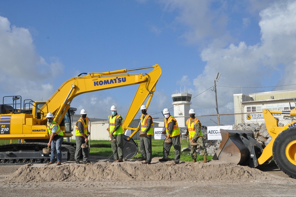 A groundbreaking ceremony is taking place at what appears to be a military airfield or installation, with several individuals in hard hats, high-visibility vests, and face masks holding shovels. A large yellow Komatsu PC210LC excavator is prominently positioned behind the group, alongside a wheel loader. AECOM signage is visible in the background, indicating contractor involvement in the project.