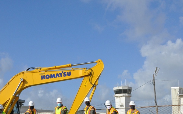 A groundbreaking ceremony is taking place at what appears to be a military airfield or installation, with several individuals in hard hats, high-visibility vests, and face masks holding shovels. A large yellow Komatsu PC210LC excavator is prominently positioned behind the group, alongside a wheel loader. AECOM signage is visible in the background, indicating contractor involvement in the project.
