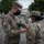 Two U.S. Army soldiers in OCP (Operational Camouflage Pattern) uniforms are engaged in what appears to be a military award or promotion ceremony outdoors. The senior soldier on the left is pinning something onto the uniform of the soldier on the right, who is identified by a name tape reading 'MCLAU[ghlin]'. Both soldiers are wearing face masks, suggesting this took place during the COVID-19 pandemic period.
