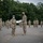 A military ceremony is taking place outdoors on a paved surface, where two soldiers in the foreground are exchanging what appears to be a certificate or commendation document. All personnel are wearing face masks, suggesting this occurred during the COVID-19 pandemic period. A formation of soldiers stands at attention in the background, spaced apart in a socially distanced arrangement.