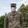 A U.S. Army soldier identified by name tape as 'KERN' stands outdoors wearing Operational Camouflage Pattern (OCP) uniform and an N95-style respirator mask. The soldier wears glasses and an OCP patrol cap with a rank insignia visible on the front. The setting appears to be an outdoor area with trees and a partly cloudy sky in the background.