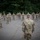A group of approximately 20-25 military personnel in camouflage uniforms are gathered in an outdoor parking lot or paved area, socially distanced and wearing face masks. One soldier stands with their back to the camera facing the group, appearing to be in a leadership or addressing role. The group is applauding, suggesting a ceremony, recognition event, or formal address.