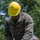 A U.S. Marine in MARPAT digital camouflage uniform is operating a chainsaw to cut through a log in an outdoor wooded area. The Marine is wearing a yellow hard hat and black protective gloves labeled 'CLASSIC' for safety. The name tape visible on the uniform reads 'FORBES' and the branch tape reads 'U.S. MARINES'.