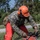 A person in U.S. Marine Corps MARPAT camouflage uniform is operating a Makita chainsaw to cut through a fallen tree or large branch. The individual is wearing full personal protective equipment including an orange hard hat, ear protection, orange face covering, tactical gloves, and orange chainsaw chaps. The scene appears to be outdoors near a road, likely during debris clearing operations.