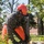 A military service member in MARPAT digital camouflage uniform is operating a Makita chainsaw to cut through a fallen tree. The individual is wearing full personal protective equipment including an orange hard hat, ear protection, face shield, orange chainsaw chaps, and black gloves. The setting appears to be a base or institutional grounds with trees and a brick building visible in the background.