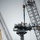 The image shows a naval vessel undergoing maintenance or construction work in a shipyard. Two workers wearing hard hats and safety harnesses are standing atop a ship's mast or superstructure platform, with a large crane lowering rigging toward them. Multiple cranes are visible surrounding the vessel, indicating a significant refit or installation operation.