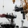 The image shows a large naval vessel undergoing construction or major refit work at a shipyard. Workers in safety gear are visible on elevated platforms and walkways around what appears to be the island superstructure of an aircraft carrier or large amphibious assault ship. A large yellow lattice crane is prominently visible on the right side, actively supporting the operation.