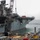 A large gray naval warship, likely an amphibious assault ship, is docked at a shipyard undergoing maintenance or repair operations. The vessel shows signs of wear including rust patches, and is surrounded by scaffolding, cranes, and industrial equipment on the pier. Workers in safety gear are visible on the dock area managing the refit operations.