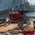 Workers in safety gear are performing maintenance or repair operations on a large naval vessel docked at a shipyard. A group of four workers are elevated in a red personnel lift basket, positioned near a structural component of the ship. Additional workers are visible on the dock below, coordinating the operation.